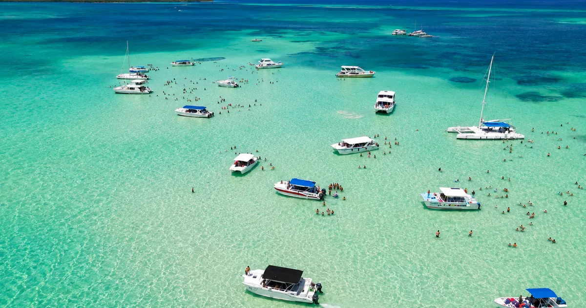 Dozens of tour boats anchored around the shallow natural pool off Saona at midday, with guests standing in the water